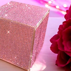 May include: A pink square box covered in sparkling rhinestones. The box is sitting on a light pink surface.
