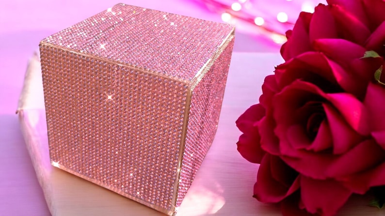May include: A pink square box covered in sparkling rhinestones. The box is sitting on a light pink surface.