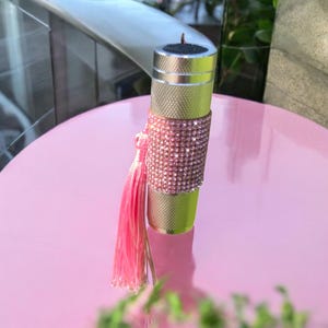 Può includere: Oggetto cilindrico dorato e strutturato, ornato da una fascia di strass rosa. Un nappa rosa è appesa. Probabilmente è un accessorio decorativo.