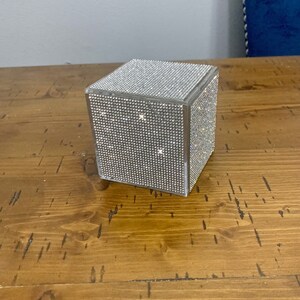 May include: A clear, square box covered in sparkling rhinestones. The box is sitting on a wooden surface.