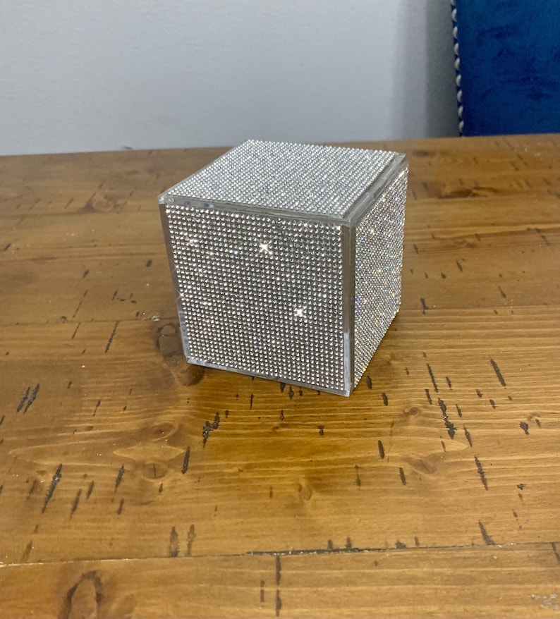 May include: A clear, square box covered in sparkling rhinestones. The box is sitting on a wooden surface.