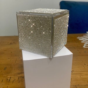 May include: A clear cube-shaped box covered in sparkling rhinestones. The box is sitting on a white box.