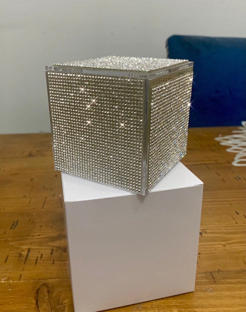 May include: A clear cube-shaped box covered in sparkling rhinestones. The box is sitting on a white box.