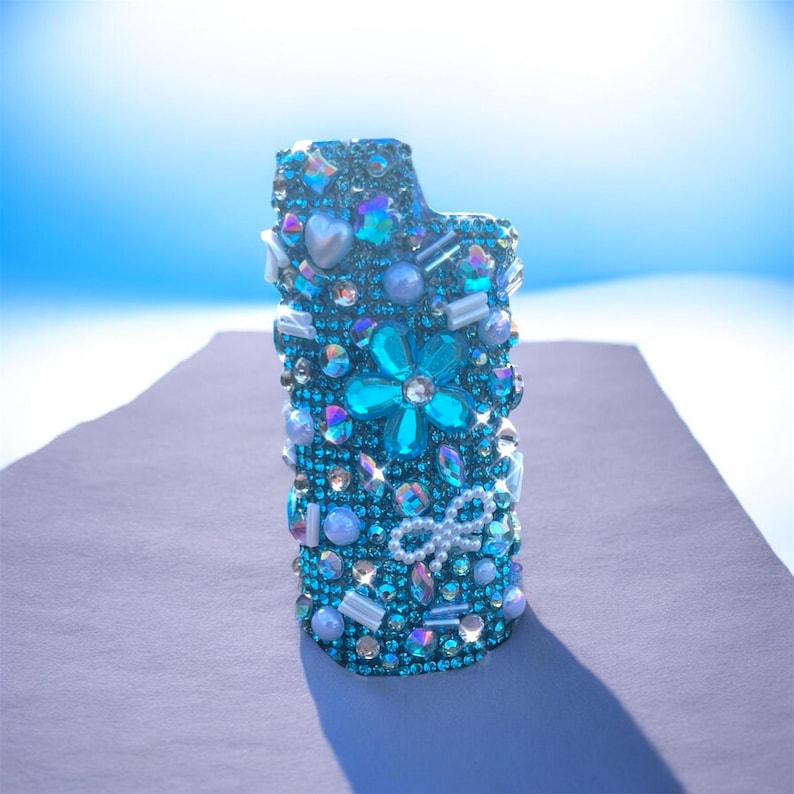Peut inclure: Un &eacute;tui de briquet bleu sarcelle orn&eacute; de bijoux. L'&eacute;tui est recouvert de strass iris&eacute;s et bleus, de perles et d'&eacute;l&eacute;ments d&eacute;coratifs de diff&eacute;rentes tailles. Une grande fleur turquoise et un n&oelig;ud blanc sont des &eacute;l&eacute;ments importants.