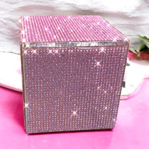以下が含まれることがあります： 正方形のピンク色のラインストーンボックス、シルバーのトリム付き。ボックスは小さく輝くラインストーンで覆われています。ボックスはピンク色の表面に置かれています。