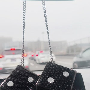 K&ouml;nnte beinhalten: Paar schwarze W&uuml;rfel-Autoanh&auml;nger, die an einer silbernen Kette h&auml;ngen. Jeder W&uuml;rfel ist mit kleinen, funkelnden schwarzen Strasssteinen mit wei&szlig;en Punktmarkierungen bedeckt. Die W&uuml;rfel h&auml;ngen vor einem Armaturenbrett.