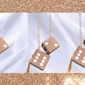 K&ouml;nnte beinhalten: Drei Paare h&auml;ngender W&uuml;rfel, jeweils mit einer goldenen Kette und Quaste. Die W&uuml;rfel sind beige mit wei&szlig;en Punkten. Der Hintergrund ist wei&szlig;er Stoff mit einem goldenen Glitzerrahmen.
