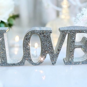 May include: A silver glitter "LOVE" sign, surrounded by white and pink roses and lit candles. The letters sparkle, creating a romantic atmosphere, ideal for weddings or Valentine's Day. The scene is elegant and soft.