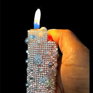 Peut inclure: Un briquet d&eacute;coratif orn&eacute; de petits strass scintillants et de fausses perles. La flamme est d'un blanc et bleu vifs, sur fond noir. Le briquet est tenu dans une main.