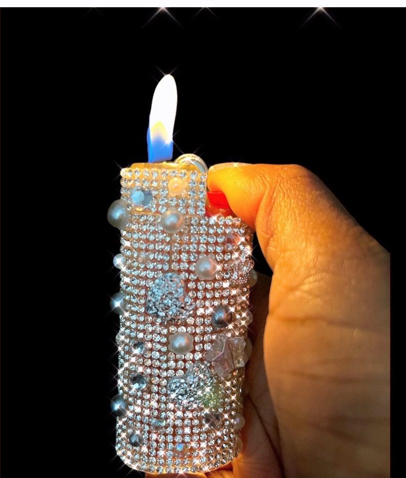 Peut inclure: Un briquet d&eacute;coratif orn&eacute; de petits strass scintillants et de fausses perles. La flamme est d'un blanc et bleu vifs, sur fond noir. Le briquet est tenu dans une main.
