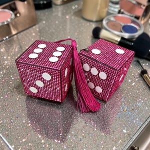 K&ouml;nnte beinhalten: Zwei rosa, strassbesetzte W&uuml;rfel mit wei&szlig;en Punkten, jeweils mit einem passenden rosa Quastenanh&auml;nger. Die W&uuml;rfel liegen auf einer glitzernden Oberfl&auml;che, mit Make-up-Pinseln und Puderdosen im Hintergrund.