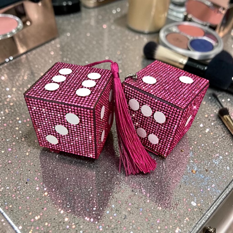 K&ouml;nnte beinhalten: Zwei rosa, strassbesetzte W&uuml;rfel mit wei&szlig;en Punkten, jeweils mit einem passenden rosa Quastenanh&auml;nger. Die W&uuml;rfel liegen auf einer glitzernden Oberfl&auml;che, mit Make-up-Pinseln und Puderdosen im Hintergrund.