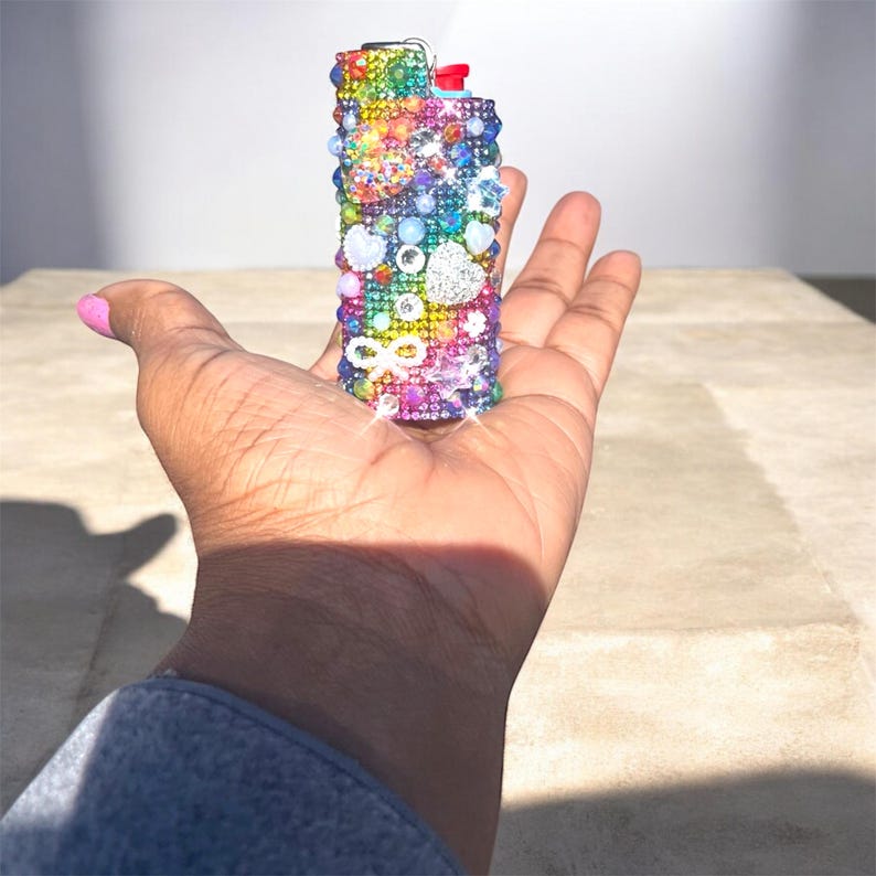 Peut inclure: Un briquet rectangulaire orn&eacute; de strass et de pierres pr&eacute;cieuses color&eacute;es. Le briquet pr&eacute;sente un arc-en-ciel de couleurs, dont le rouge, l'orange, le jaune, le vert, le bleu et le violet. Des &eacute;l&eacute;ments d&eacute;coratifs incluent des c&oelig;urs, des &eacute;toiles et un symbole infini.