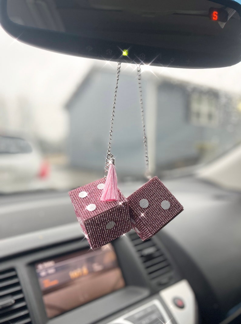 K&ouml;nnte beinhalten: Rosafarbene W&uuml;rfel aus Strasssteinen, die an einem R&uuml;ckspiegel h&auml;ngen. Die W&uuml;rfel sind mit kleinen, glitzernden Strasssteinen besetzt und haben wei&szlig;e Punkte. Zwischen den W&uuml;rfeln h&auml;ngt eine rosa Quaste.