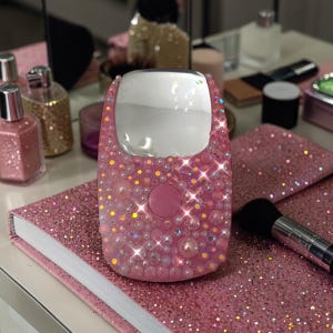 May include: A pink, bedazzled handheld mirror with a large, rectangular mirror surface. The mirror is covered in small, sparkling crystals and pearls. It rests on a pink, glittery notebook, with a makeup brush nearby. Other beauty products are in the background.