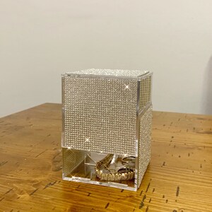 May include: A clear acrylic jewelry organizer with a diamond-like pattern on the exterior. The organizer has multiple compartments for storing rings, bracelets, and other jewelry. The organizer is sitting on a wooden surface.