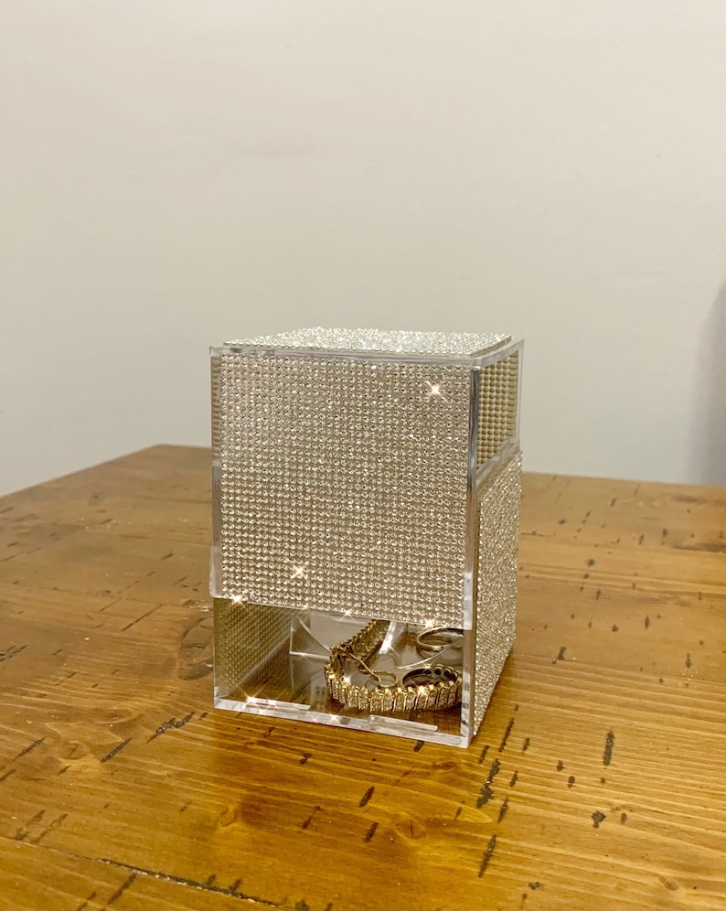 May include: A clear acrylic jewelry organizer with a diamond-like pattern on the exterior. The organizer has multiple compartments for storing rings, bracelets, and other jewelry. The organizer is sitting on a wooden surface.
