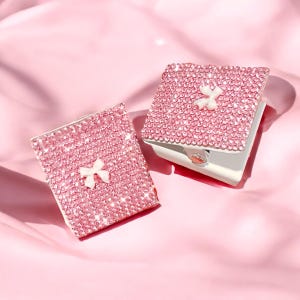 May include: Two pink, bedazzled, square-shaped compact mirrors with white bows. The mirrors are covered in small, sparkling pink rhinestones. The compacts are on a pink satin surface.