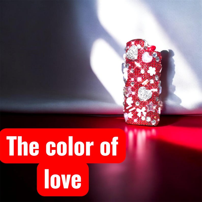 Peut inclure: Un briquet rouge orn&eacute; de strass, de c&oelig;urs et de motifs floraux. Le briquet a un petit n&oelig;ud rouge et le texte "The color of love" dans un rectangle rouge.