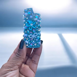 Peut inclure: Un &eacute;tui de briquet bleu orn&eacute; de strass iris&eacute;s, de perles et d'un motif floral. L'&eacute;tui pr&eacute;sente un n&oelig;ud et diverses formes d'ornements bleus et transparents. L'&eacute;tui du briquet est tenu dans une main avec du vernis &agrave; ongles noir.