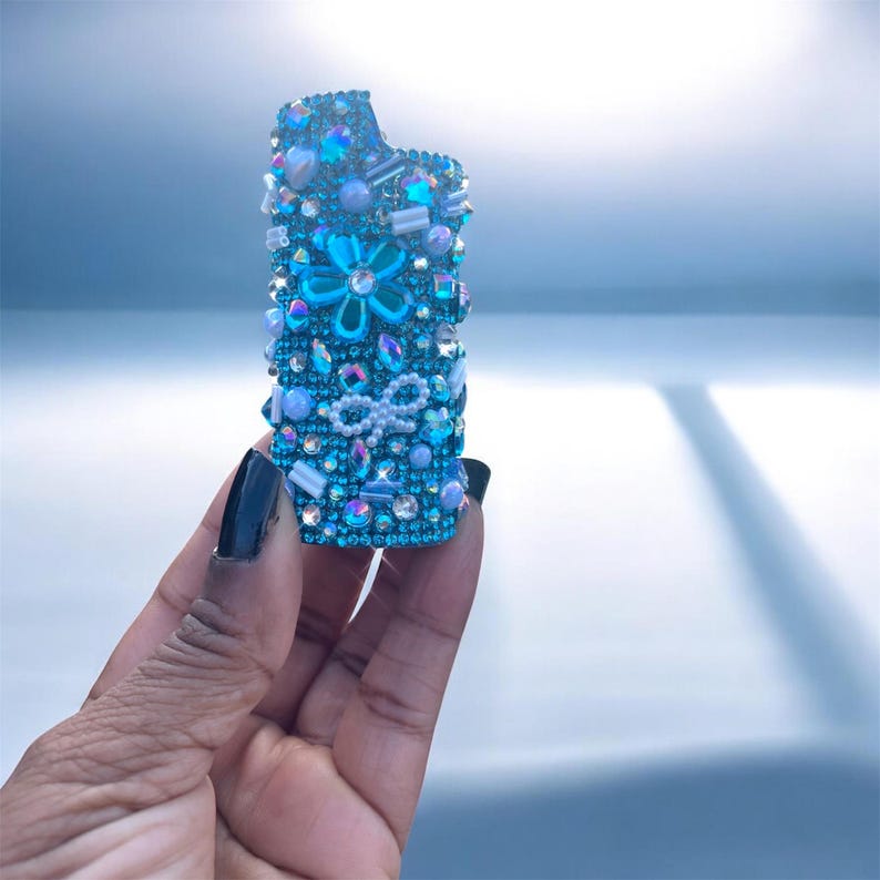 Peut inclure: Un &eacute;tui de briquet bleu orn&eacute; de strass iris&eacute;s, de perles et d'un motif floral. L'&eacute;tui pr&eacute;sente un n&oelig;ud et diverses formes d'ornements bleus et transparents. L'&eacute;tui du briquet est tenu dans une main avec du vernis &agrave; ongles noir.