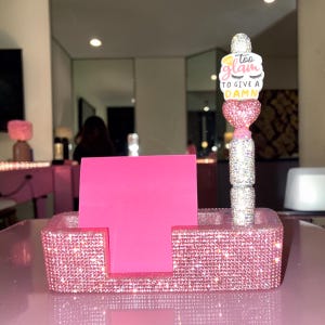 Op de afbeelding: Een rechthoekige bureau-organizer bedekt met roze strass-steentjes, met een stapel felroze plakbriefjes en een bijpassende strass-pen. De pen is bekroond met een wit hartvormig sieraad met de tekst "too glam TO GIVE A DAMN".