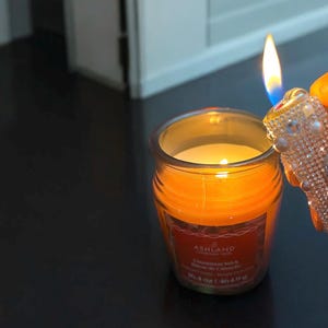 Peut inclure: Une bougie allum&eacute;e, de couleur orange, dans un pot en verre avec une &eacute;tiquette qui dit "Ashland Cinnamon Stick". Un briquet orn&eacute; est tenu pr&egrave;s de la flamme de la bougie. La bougie est de 113 g.