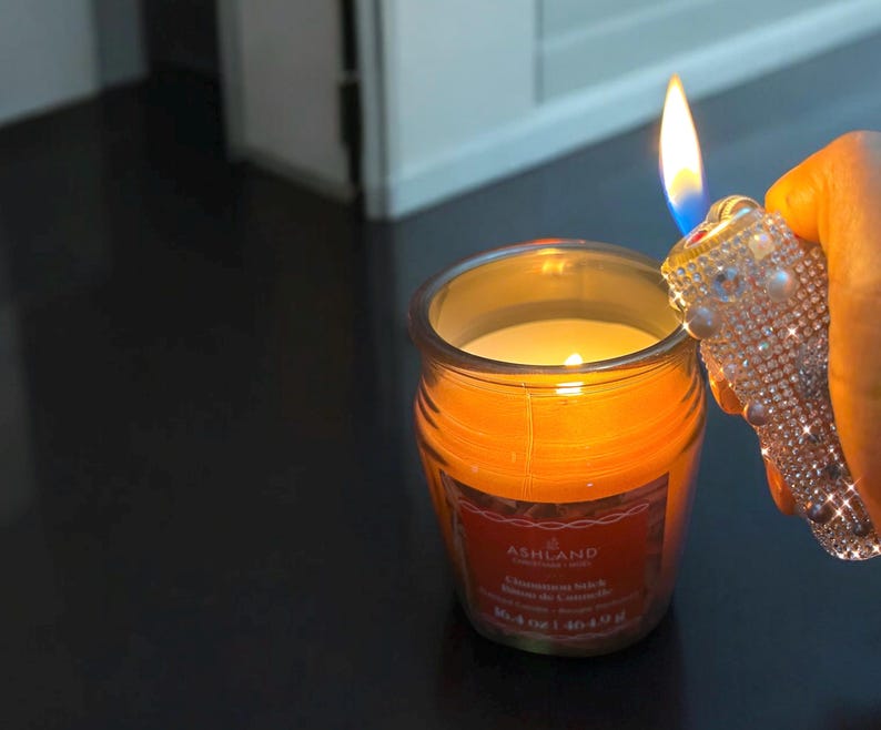 Peut inclure: Une bougie allum&eacute;e, de couleur orange, dans un pot en verre avec une &eacute;tiquette qui dit "Ashland Cinnamon Stick". Un briquet orn&eacute; est tenu pr&egrave;s de la flamme de la bougie. La bougie est de 113 g.