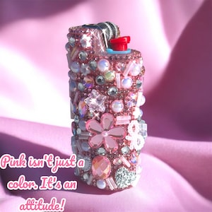 Peut inclure: Un briquet rose orn&eacute; de strass, perles et pierres pr&eacute;cieuses de diff&eacute;rentes tailles et formes. Le briquet a un bouton rouge et un dessus en m&eacute;tal noir et argent&eacute;. Le texte de l'image se lit comme suit : "Le rose n'est pas qu'une couleur. C'est une attitude !"
