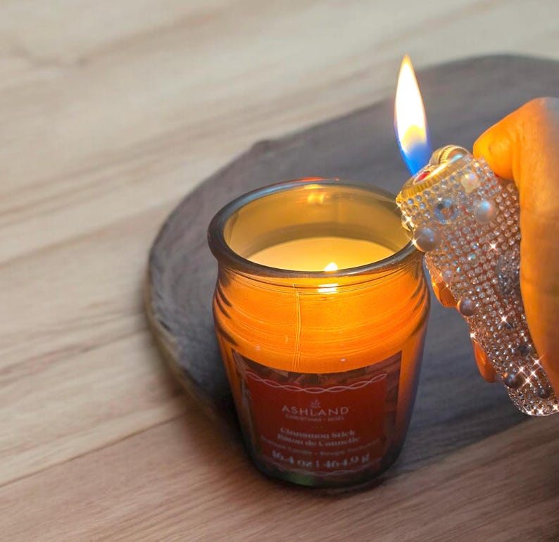 Peut inclure: Une bougie allum&eacute;e dans un pot en verre avec une &eacute;tiquette rouge indiquant "Ashland Cinnamon Stick". Un briquet d&eacute;coratif avec des strass est tenu pr&egrave;s de la flamme. La bougie est pos&eacute;e sur une surface en bois.