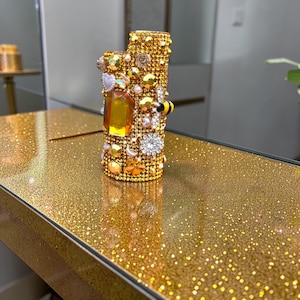 Peut inclure: Un briquet dor&eacute; incrust&eacute; de diverses pierres pr&eacute;cieuses, perles et &eacute;l&eacute;ments d&eacute;coratifs. Le briquet pr&eacute;sente une grosse gemme ambr&eacute;e et une petite abeille d&eacute;corative. La surface est recouverte de paillettes dor&eacute;es.