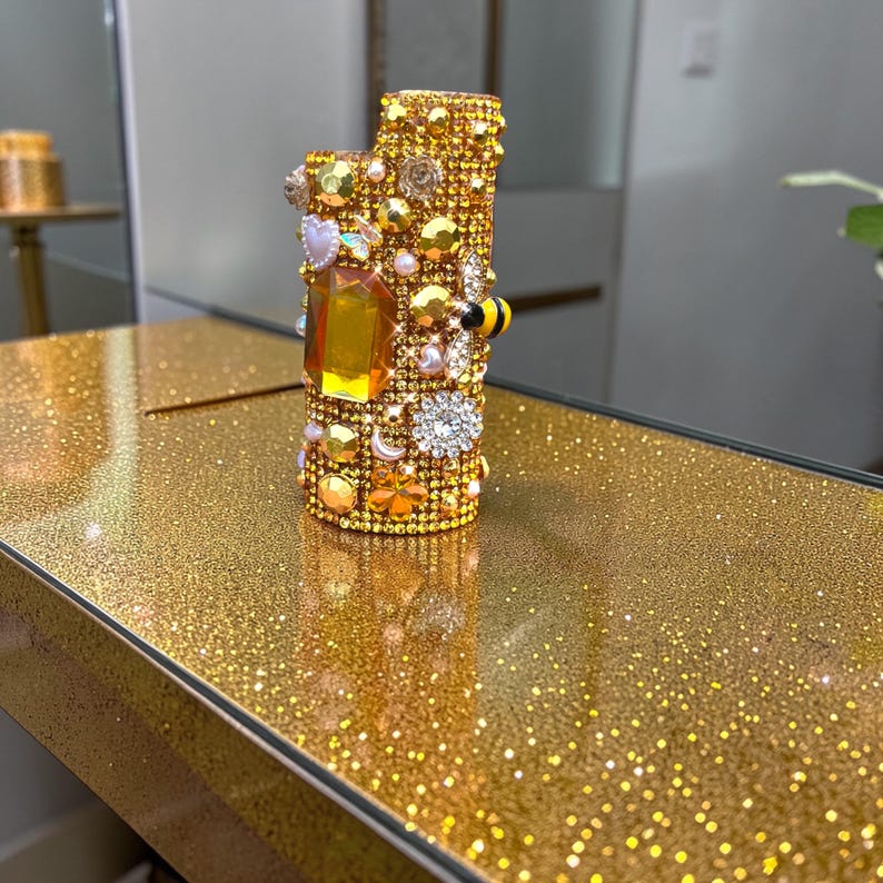 Peut inclure: Un briquet dor&eacute; incrust&eacute; de diverses pierres pr&eacute;cieuses, perles et &eacute;l&eacute;ments d&eacute;coratifs. Le briquet pr&eacute;sente une grosse gemme ambr&eacute;e et une petite abeille d&eacute;corative. La surface est recouverte de paillettes dor&eacute;es.