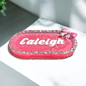 May include: A rectangular, pink resin tray with the name "Caleigh" in white script. The tray is edged with silver glitter and adorned with a pink bow with a silver gem center. A decorative item.