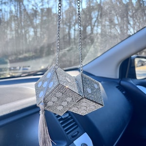 K&ouml;nnte beinhalten: Ein Auto-Accessoire mit zwei W&uuml;rfeln, die jeweils mit glitzernden Strasssteinen und wei&szlig;en Punkten bedeckt sind. Die W&uuml;rfel sind verbunden und h&auml;ngen an einer silbernen Kette, mit einer silbernen Quaste, die an einem W&uuml;rfel h&auml;ngt. Der Innenraum eines Autos ist im Hintergrund zu sehen.