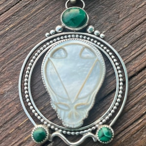 May include: A silver pendant necklace with a white shell skull design, surrounded by a beaded circle and accented with three green malachite stones.