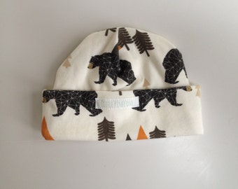 May include: A cream-colored beanie hat with a repeating pattern of black bears, brown trees, and orange triangles. The hat has a folded brim and the text "LITTLE PINECONE" is visible on the brim.
