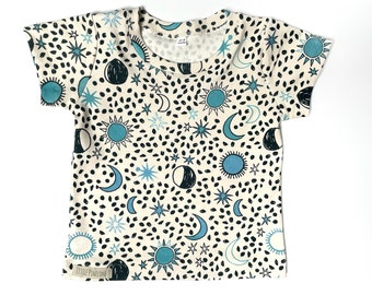 May include: Cream-colored short-sleeved t-shirt with a celestial pattern. The design features teal suns, moons, and stars, along with black speckles. The shirt has a round neckline and a tag that reads "LITTLE PINECOVE".