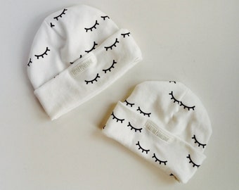 May include: Two white baby beanies with a repeating black eyelash pattern. Each beanie has a folded brim and a small label that reads "LITTLE PINEONE". The beanies are made of a soft material.