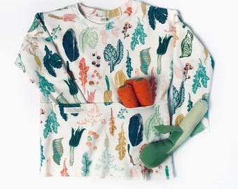 May include: White long-sleeved shirt with a colorful vegetable pattern. The shirt has a pocket with two orange carrots and a green leek. The shirt is decorated with illustrations of various vegetables and plants.