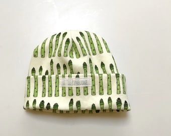 May include: A white baby hat with a green asparagus pattern. The hat has a white label with the text "Little Pinecone" on it.