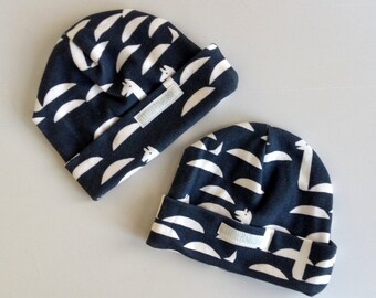May include: Two navy blue baby beanies with a white crescent and giraffe pattern. Each beanie has a folded brim and a small label. The beanies are made of soft fabric.