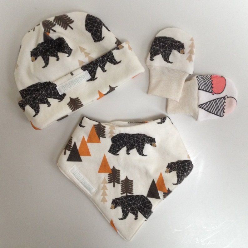 May include: A baby set featuring a cream-colored beanie, bib, and mittens. The set is patterned with black bears, brown trees, and orange triangles. The beanie has a folded brim, and the bib has a velcro closure.