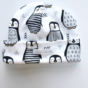 May include: White beanie featuring a repeating pattern of black and white penguins with closed eyes. The hat has a folded brim and small gray triangles. The text "LITTLE PINECONE" is on a small label.