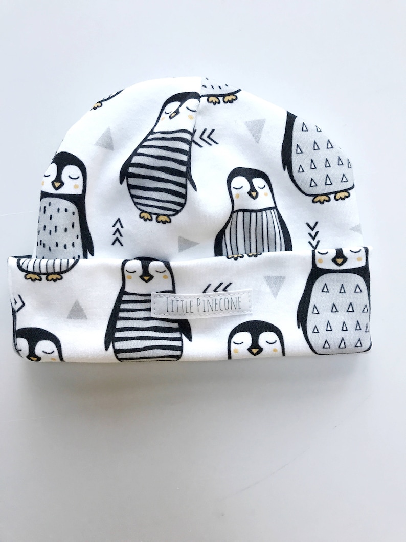 May include: White beanie featuring a repeating pattern of black and white penguins with closed eyes. The hat has a folded brim and small gray triangles. The text "LITTLE PINECONE" is on a small label.