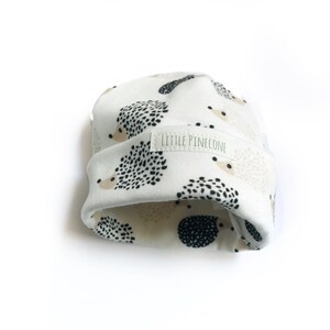 May include: White baby hat with a black and beige hedgehog print. The hat has a label that says "Little Pinecone".