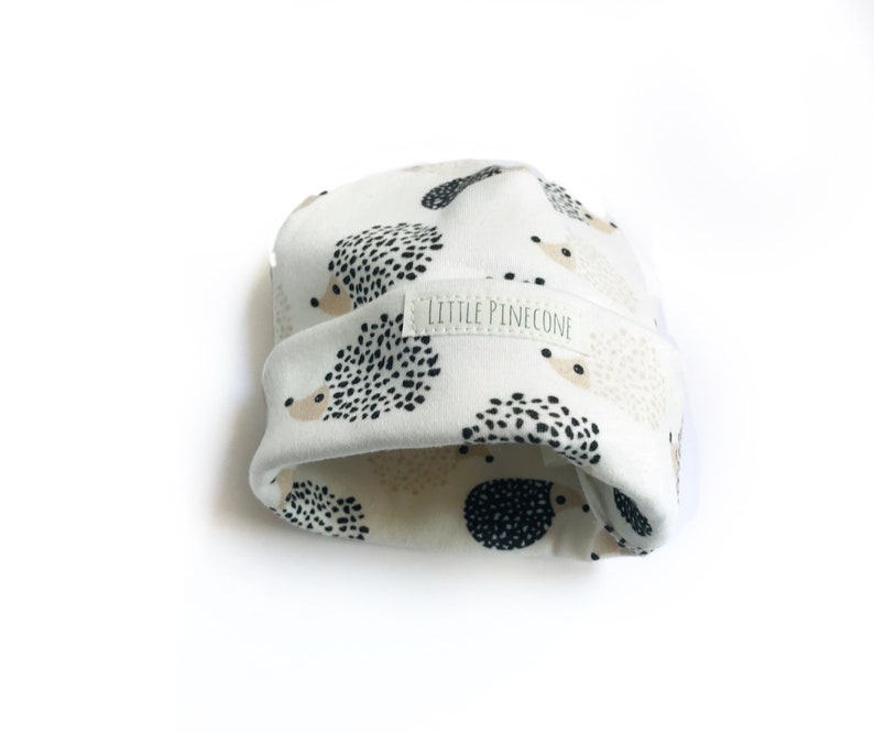 May include: White baby hat with a black and beige hedgehog print. The hat has a label that says "Little Pinecone".