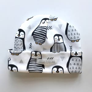 May include: White beanie featuring a repeating pattern of cartoon penguins in black and white. The penguins have various designs, including stripes and triangles. The hat has a folded brim and a small label that reads "LITTLE PINECONE".
