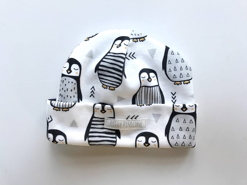 May include: White beanie featuring a repeating pattern of cartoon penguins in black and white. The penguins have various designs, including stripes and triangles. The hat has a folded brim and a small label that reads "LITTLE PINECONE".