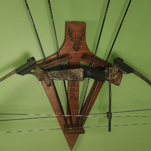 May include: A wooden bow and arrow rack with a deer head design. The rack is mounted on a green wall and has a bow and five arrows attached to it. The rack is labeled "muci".