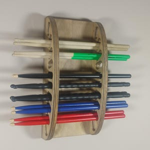 May include: Wooden wall-mounted drum stick holder with multiple slots for storing drumsticks. The holder has a natural wood finish and is filled with drumsticks in various colors, including red, blue, black, and green.